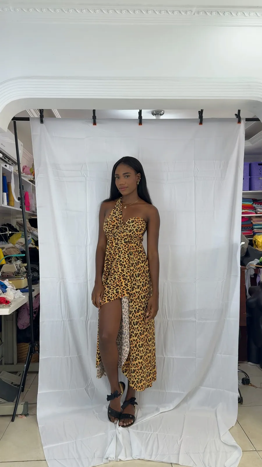 Toma original en el taller: la modelo con el Vestido Brisa en leopardo frente a una sábana blanca, con estantes de telas alrededor